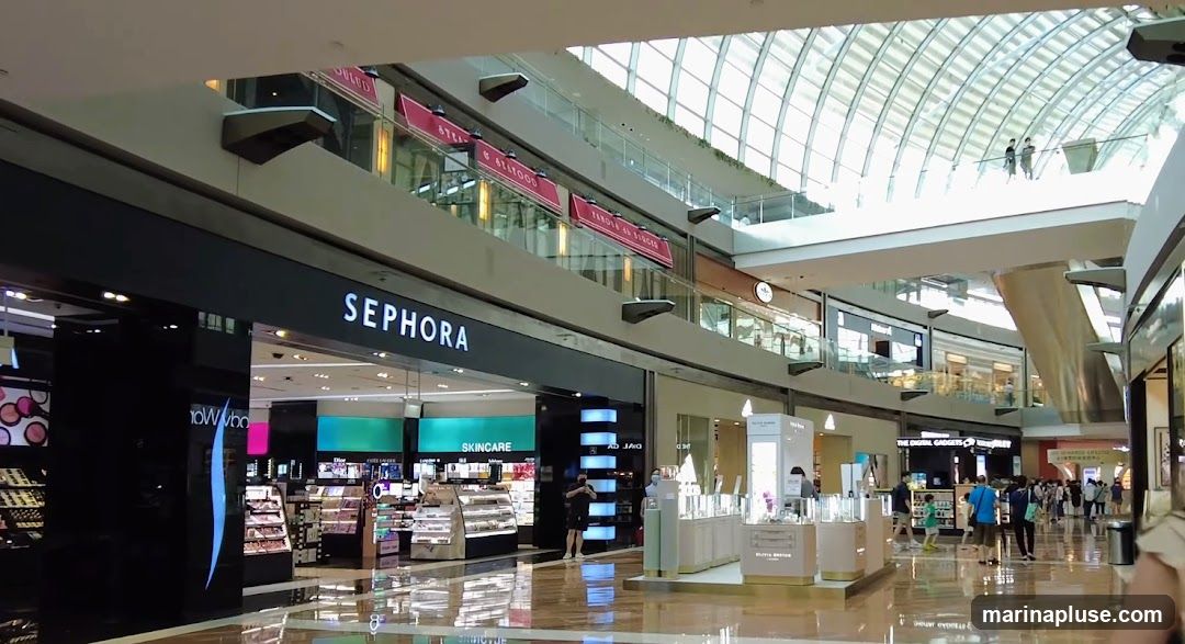 A wide shot of a Sephora store entrance within a modern, multi-level shopping mall. The Sephora sign is prominently displayed above the entrance. Inside the store, various cosmetics and skincare products are displayed on shelves and in illuminated cases. The mall features a large, arched glass ceiling, allowing natural light to illuminate the interior. Other stores and shoppers are visible in the background, along with escalators and walkways connecting different levels.