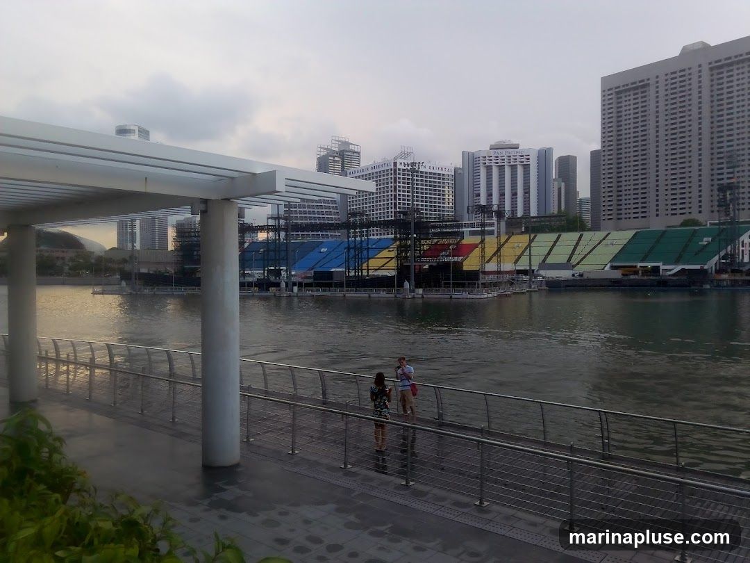 A view of a waterfront promenade with a modern canopy structure in the foreground. In the background, across the water, is a large stadium with colorful tiered seating. Several tall buildings form the city skyline behind the stadium. Two people are visible on the promenade, looking out at the water. The sky is overcast.
