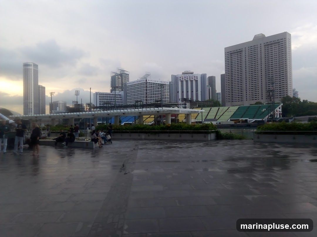 A wide shot of a cityscape with several tall buildings and a stadium in the background. The foreground features a wet, tiled plaza with a few people sitting and standing. The sky is overcast, suggesting it might have recently rained. The stadium has green and yellow seating. Signs for 'PAN PACIFIC' and 'MANDARIN ORIENTAL' are visible on buildings.