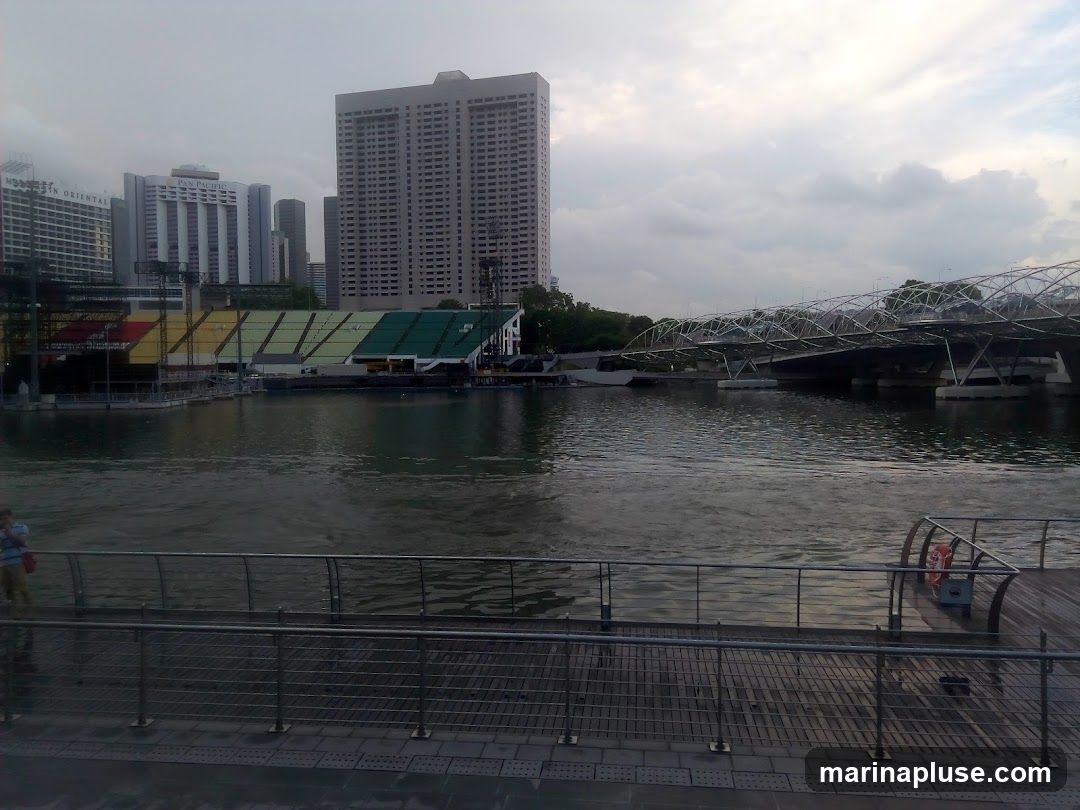 A wide shot of Marina Bay in Singapore, featuring the iconic Helix Bridge and a stadium with colorful seating. Tall buildings, including the Pan Pacific and Marina Mandarin hotels, form the city skyline in the background. The water is calm, reflecting the overcast sky. A person stands on a walkway in the foreground.