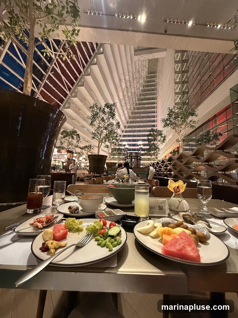 A table set with plates of partially eaten food, including fruit salad, green salad, and what appears to be some sort of appetizer. The background shows a large, modern atrium with tiered seating and decorative elements.
