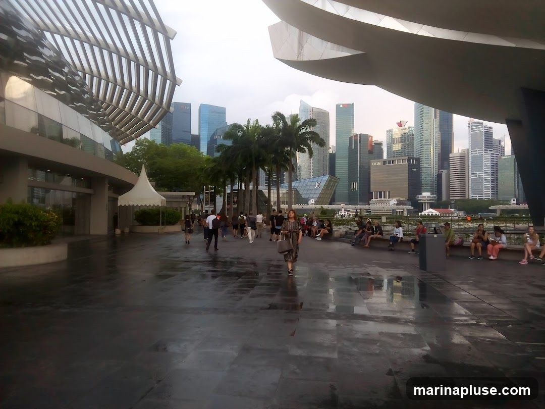 A group of people are walking and sitting in a paved outdoor plaza. The plaza has a wet, reflective surface, suggesting it may have recently rained. In the background, a dense city skyline with numerous skyscrapers is visible, along with palm trees. The architecture of the foreground suggests a modern public space, possibly a tourist attraction or a commercial area.