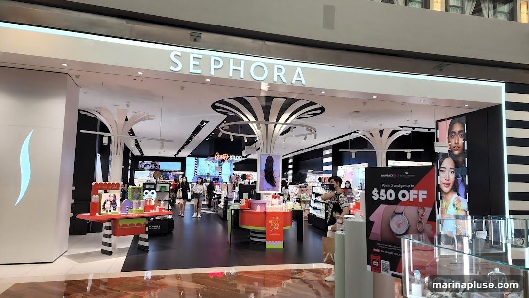The exterior of a Sephora store with the brand name prominently displayed above the entrance. Inside, the store is brightly lit and stocked with various beauty products. Displays feature makeup, skincare, and accessories. Several customers are browsing the merchandise. A large promotional poster for a ShopBack offer is visible near the entrance, advertising "$50 OFF".