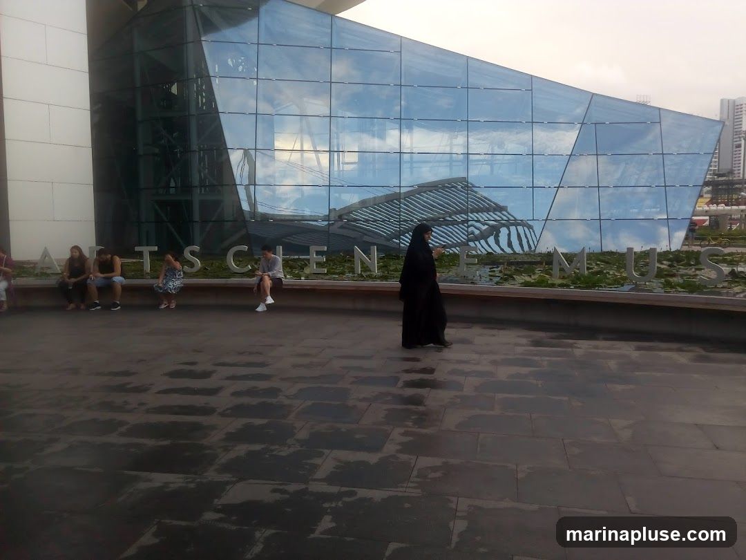 A modern building with a glass facade reflecting the sky. Large metal letters spelling out "ARTS SCIENCE MUSEUM" are visible in front of the building. Several people are sitting on a bench in front of the letters.