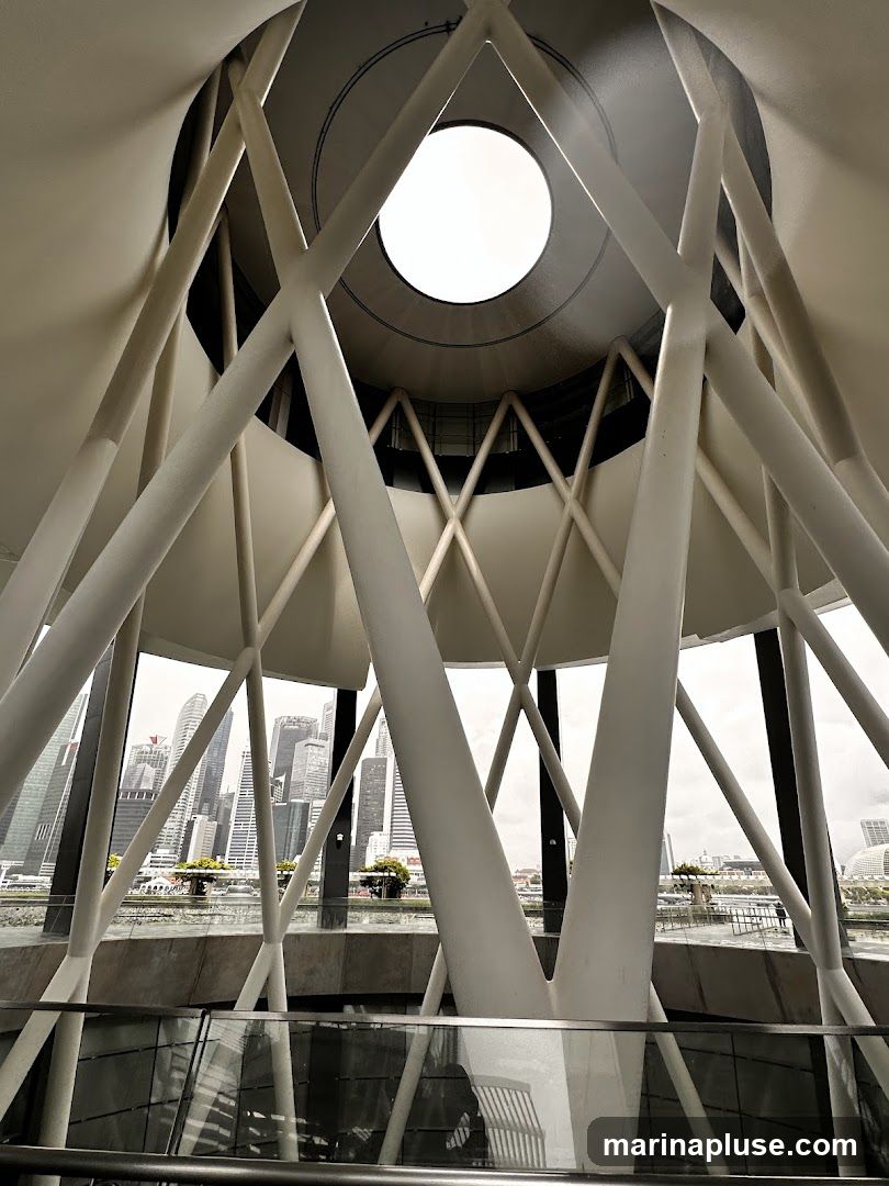 An interior view looking upwards towards a circular opening in the ceiling, framed by a complex white lattice structure. The structure's beams create a dynamic, geometric pattern. Through the openings in the structure, a panoramic view of a modern cityscape with numerous skyscrapers is visible under an overcast sky. A body of water and some greenery are also present in the mid-ground.