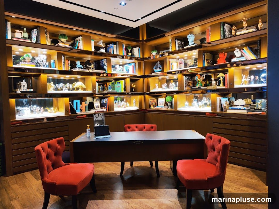 The image shows a room with extensive display shelves filled with books, decorative items, and illuminated display cases containing what appear to be watches or jewelry. In the foreground, there is a desk with a laptop and two red upholstered chairs. The overall ambiance suggests a retail or showroom setting, possibly for luxury goods.