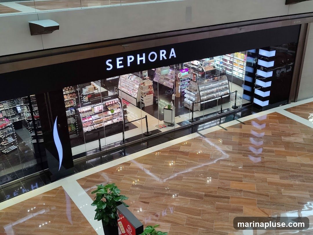 The exterior of a Sephora store is shown, with the brand name prominently displayed above the entrance. The store features large glass windows displaying various beauty products on shelves and display cases. The interior is brightly lit, showcasing a wide selection of makeup, skincare, and fragrance items. The floor in front of the store is made of polished wood-like tiles.