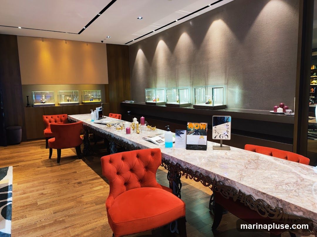 A well-lit room with a long marble-topped table adorned with decorative items and brochures. Several red velvet chairs are placed around the table. Display cases showcasing items are visible along the back wall, illuminated by spotlights. The floor is polished wood.