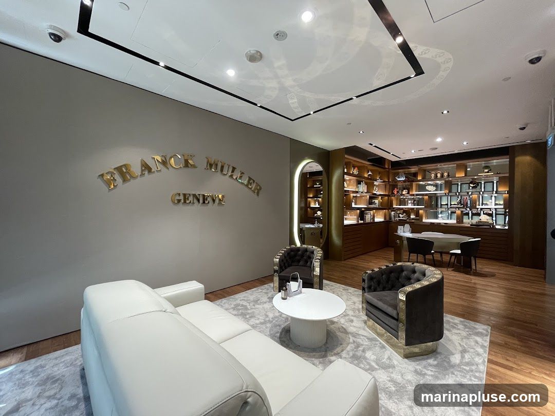 A luxurious interior of a Franck Muller store, featuring a large white sofa, two plush armchairs, and a round coffee table on a patterned rug. The walls are adorned with the brand's name, "FRANCK MULLER GENEVE," in prominent gold lettering. In the background, display shelves showcase various watches and accessories, with a consultation desk and chairs visible.