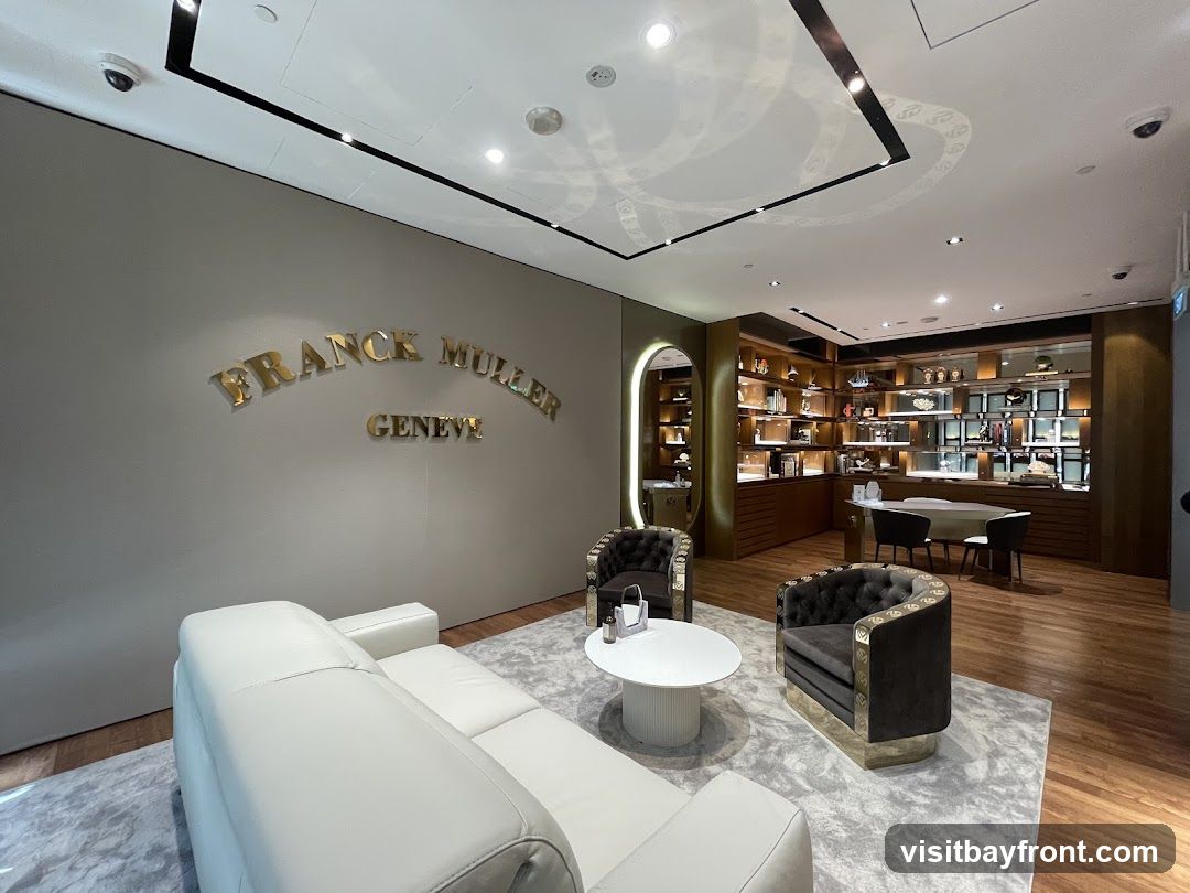 A luxurious interior of a Franck Muller store, featuring a large white sofa, two plush armchairs, and a round coffee table on a patterned rug. The walls are adorned with the brand's name, "FRANCK MULLER GENEVE," in prominent gold lettering. In the background, display shelves showcase various watches and accessories, with a consultation desk and chairs visible.