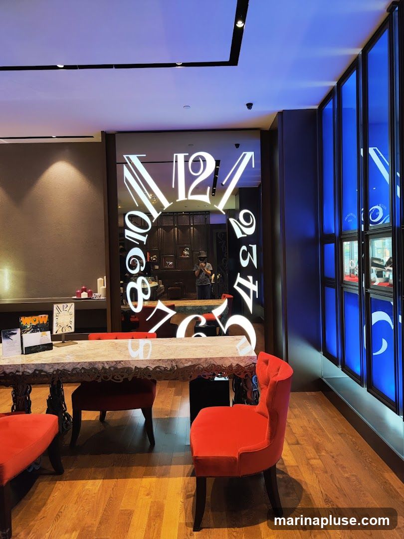 The interior of a high-end watch store features a large mirror with a clock face design, a marble-topped table with ornate legs, and plush red chairs. Display cases with blue lighting line the right wall, showcasing watches. The floor is made of polished wood.