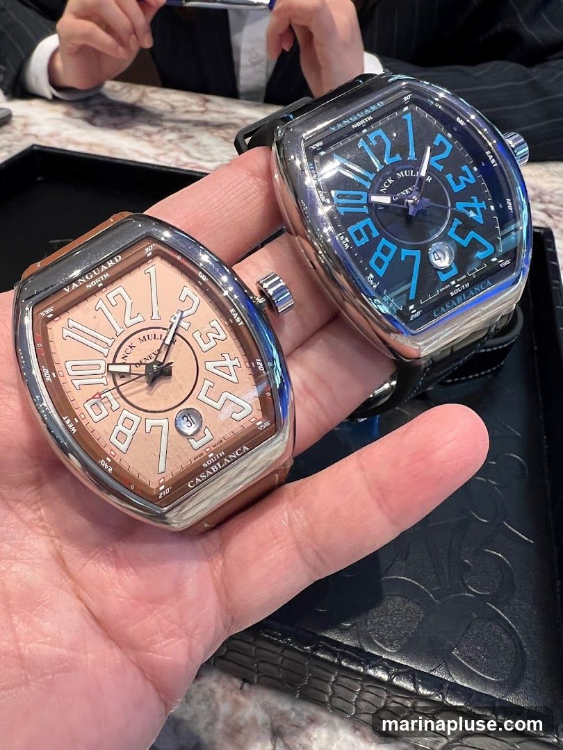 A close-up shot of two Franck Muller Vanguard Casablanca watches being held. The watch in the foreground has a light brown dial with large Arabic numerals and a date window. The watch in the background has a black dial with bright blue Arabic numerals and a date window. Both watches have the Franck Muller logo and "Vanguard Casablanca" visible on their dials. The watches are presented on a dark textured surface, possibly a watch display tray. In the background, out of focus, are people in formal attire, suggesting a retail or private viewing setting.