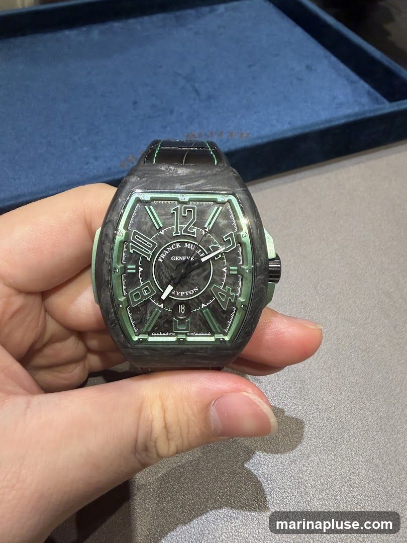 A hand holds a Franck Muller Vanguard watch with a carbon fiber case and green accents. The watch face has Arabic numerals and a date window. A blue velvet tray is visible in the background.