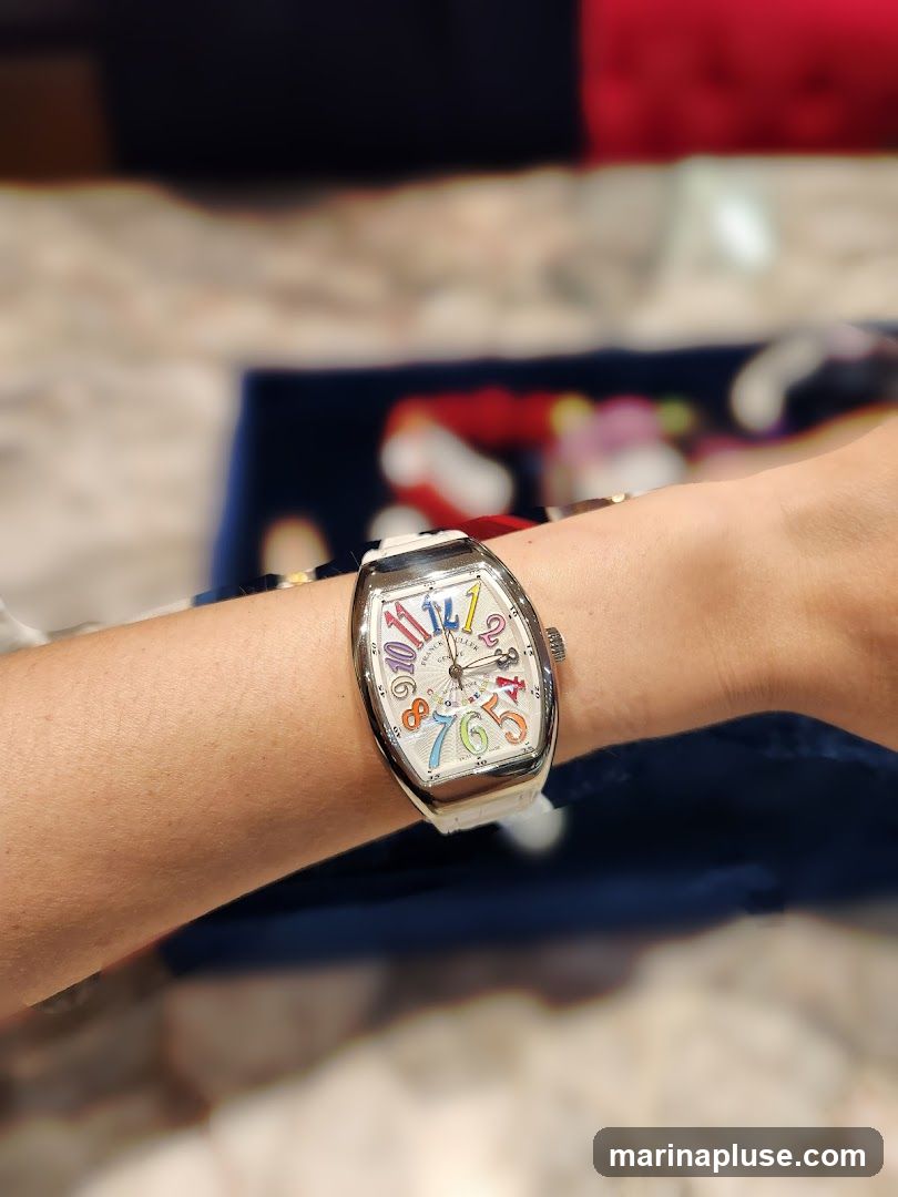 A close-up shot of a person's wrist wearing a Franck Muller Vanguard watch. The watch has a white dial with colorful Arabic numerals and a white leather strap. The background is blurred, showing hints of fabric and possibly a display case.