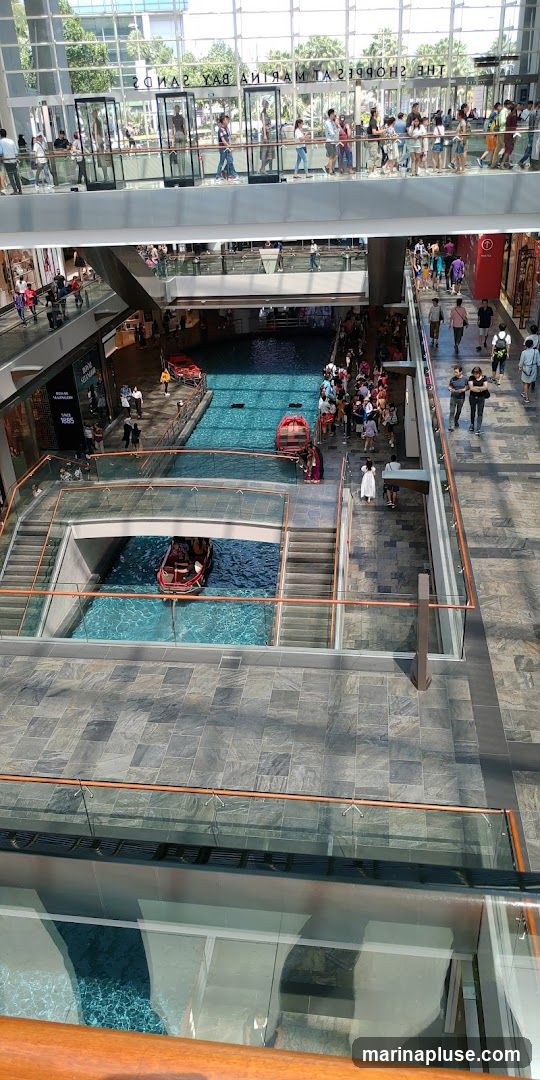 An interior view of a multi-level shopping mall with a canal running through it. People are seen walking on the upper levels and on bridges crossing the canal. Boats are present in the canal. The architecture features glass railings and modern design.