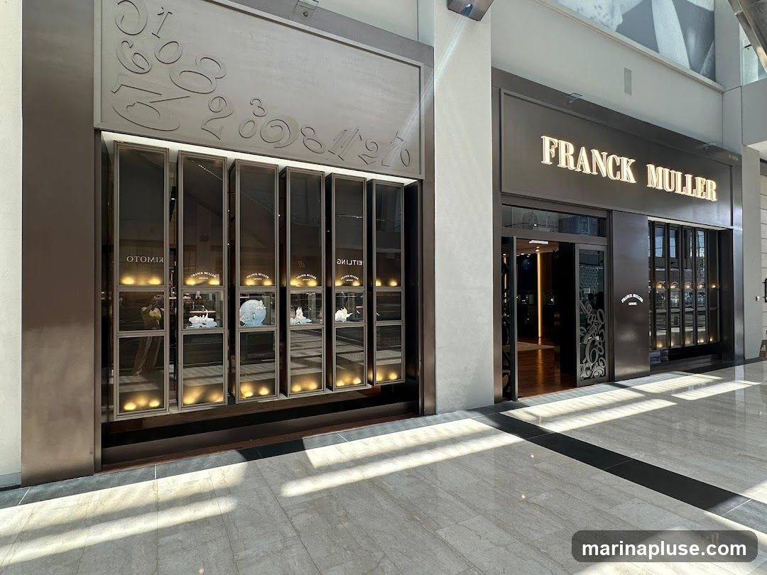 The exterior of a Franck Muller watch store. The store has a large illuminated sign that reads "FRANCK MULLER" above the entrance. To the left of the entrance is a display case with multiple compartments, each showcasing a watch or decorative item. The display case has a metallic frame and is lit from within. The store is located in a modern indoor shopping area with marble flooring.