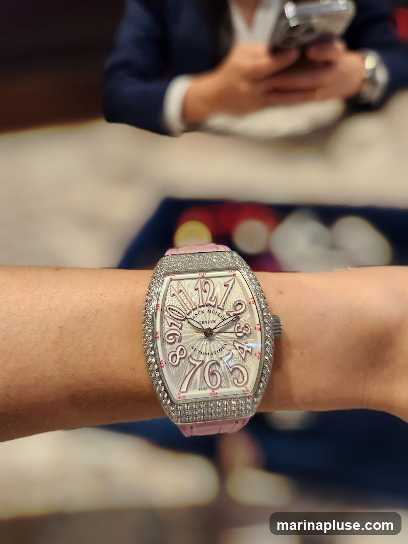 A close-up shot of a diamond-encrusted Franck Muller watch with a pink strap worn on a wrist. The watch face is silver with pink Arabic numerals and the brand name "FRANCK MULLER GENEVE" is visible. The background is blurred, showing a person in a dark suit holding a phone.