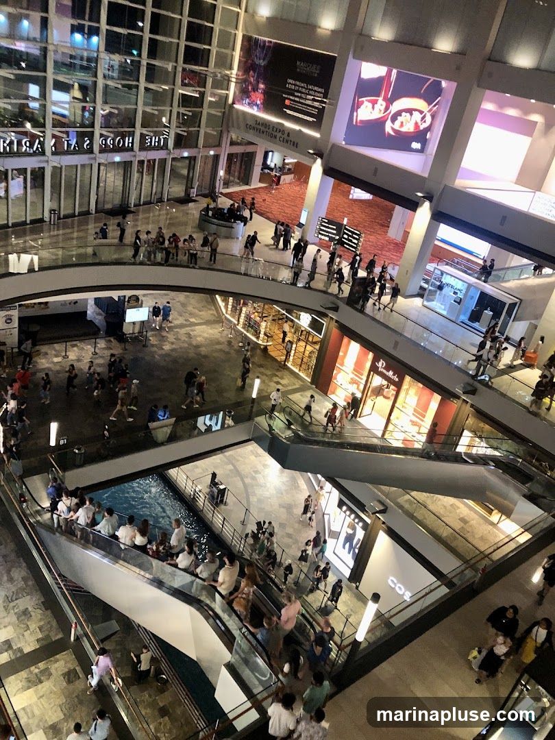 An aerial view of a multi-level shopping mall with escalators, shops, and a water feature. People are seen walking, shopping, and riding escalators. Large digital screens display advertisements and information.