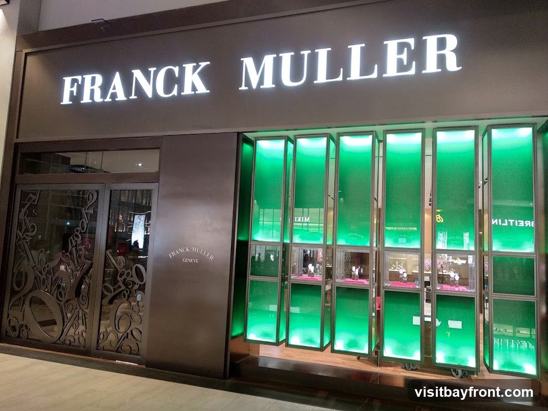The exterior of a Franck Muller watch store. The store has a large illuminated sign that reads "FRANCK MULLER" above the entrance. To the right of the entrance are several display windows illuminated with green light, showcasing watches. The entrance doors are made of dark metal with decorative patterns. A smaller "FRANCK MULLER GENEVE" logo is visible on the wall to the right of the entrance.