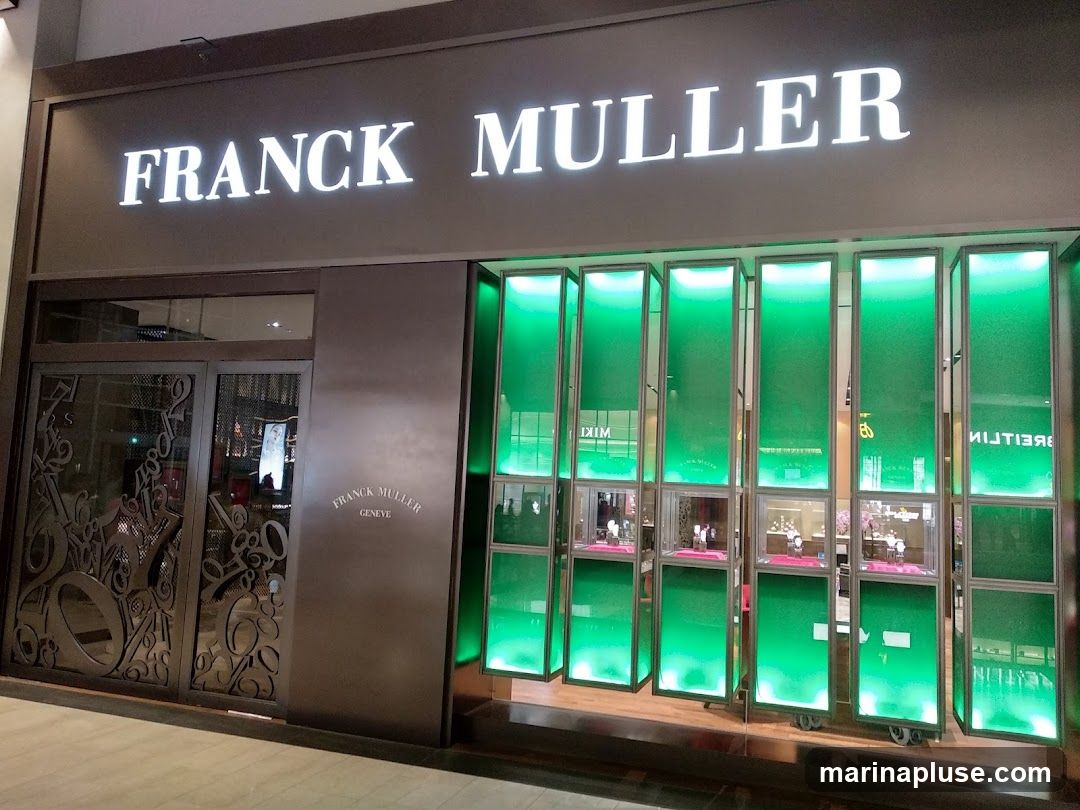The exterior of a Franck Muller watch store. The store has a large illuminated sign that reads "FRANCK MULLER" above the entrance. To the right of the entrance are several display windows illuminated with green light, showcasing watches. The entrance doors are made of dark metal with decorative patterns. A smaller "FRANCK MULLER GENEVE" logo is visible on the wall to the right of the entrance.