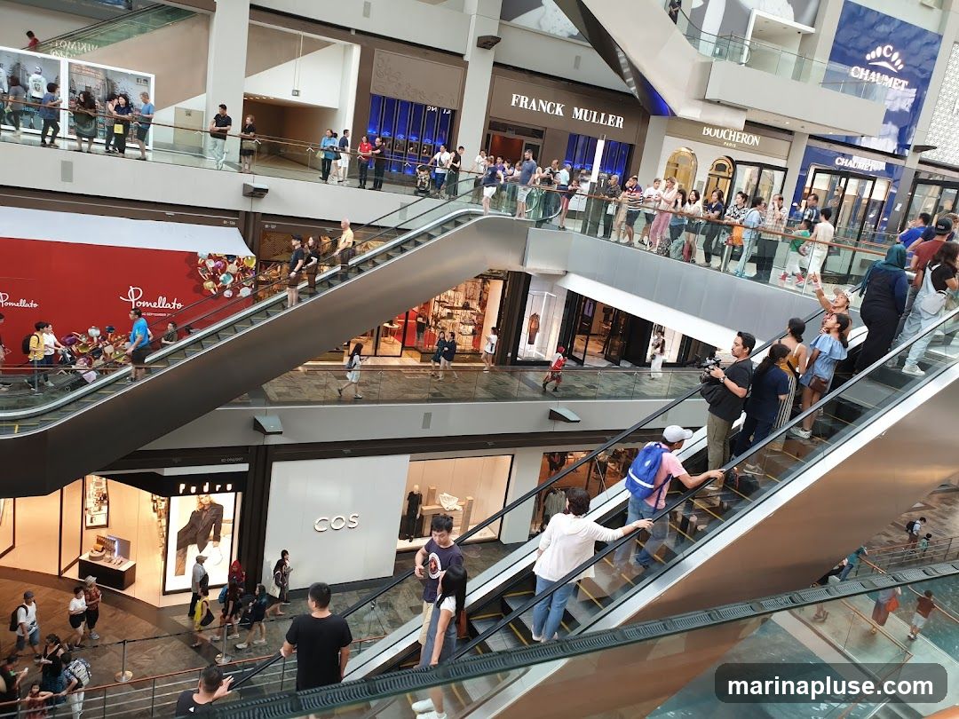 A multi-level shopping mall interior with escalators and many shoppers. Various store fronts are visible, including Franck Muller, Boucheron, Chaumet, Pomellato, and COS. People are seen walking, riding escalators, and browsing in the stores.