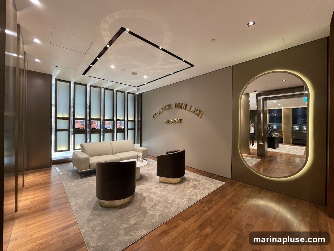 A luxurious showroom with a beige wall featuring the Franck Muller Geneve logo in gold lettering. In the foreground, a light-colored sofa and two dark armchairs are arranged on a grey rug. To the right, an oval-shaped mirror with a glowing border reflects the interior, showing display cases and a person standing near them. The floor is polished wood.