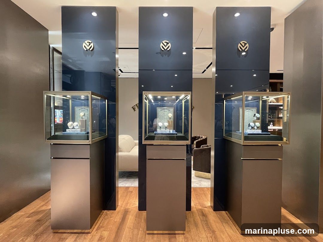 Three tall, dark blue display pillars stand in a showroom, each topped with a gold logo. Below each pillar, a glass-encased display case showcases watches on pedestals. The floor is made of polished wood. In the background, a sofa and chairs are visible, suggesting a lounge area.