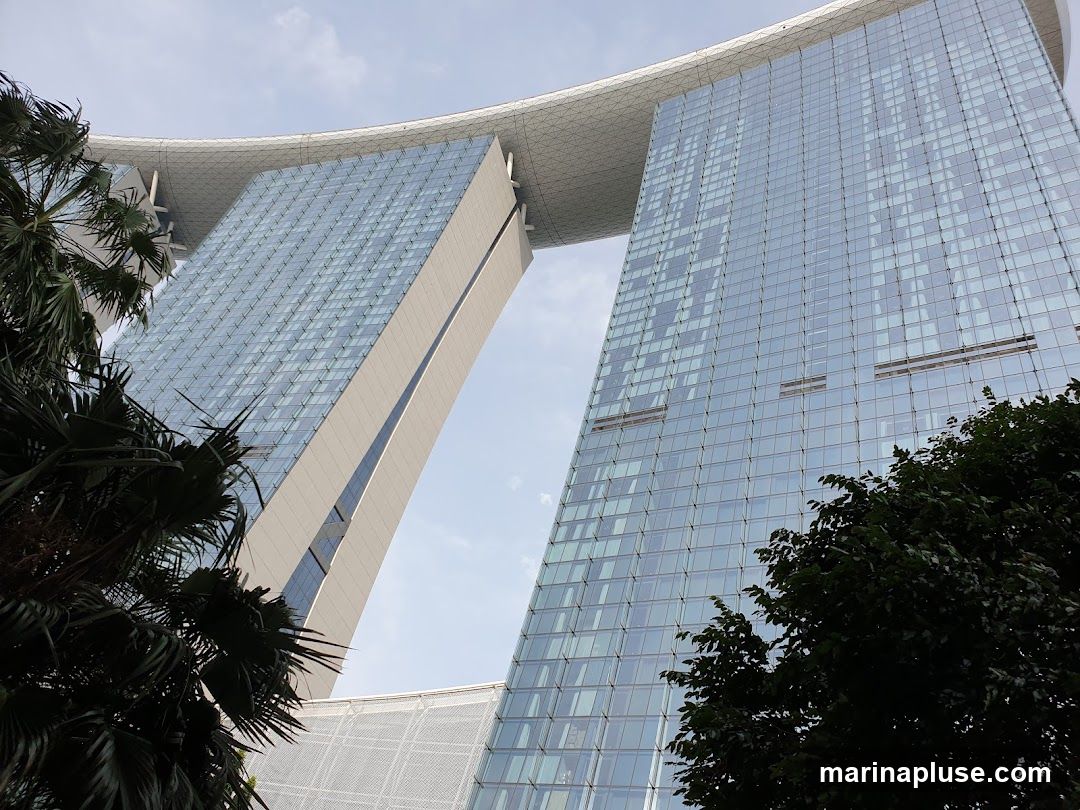 A low-angle shot of the iconic Marina Bay Sands hotel in Singapore, with its distinctive three towers connected by a sky-high platform. The glass facades of the towers reflect the cloudy sky. Lush palm trees frame the left and right sides of the image, adding a tropical element to the urban landscape.