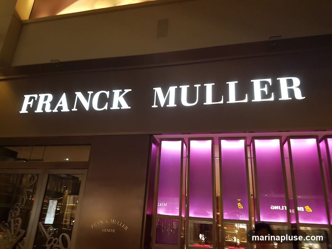 A well-lit storefront with the prominent white lettering "FRANCK MULLER" above the entrance. Below the main sign, smaller text reads "FRANCK MULLER GENEVE". The display windows are illuminated with a vibrant purple light, showcasing what appears to be watches or jewelry inside. A person is partially visible in the reflection of the window.