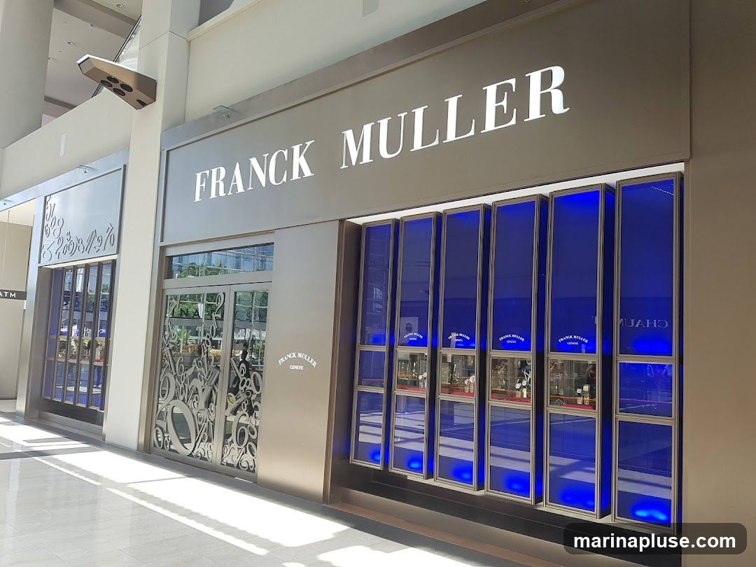 The exterior of a Franck Muller watch store. The store has a large sign above the entrance that reads "FRANCK MULLER". The storefront features several display windows with blue backlighting, showcasing watches. The entrance doors are made of glass with decorative metalwork.