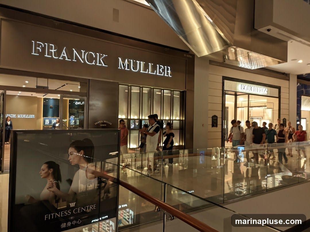 The image shows the storefronts of two luxury watch and jewelry stores, Franck Muller and Boucheron, located in a mall. The Franck Muller store has a large illuminated sign with the brand name. Inside, staff can be seen attending to customers. Adjacent to it is the Boucheron store, also with its name displayed above the entrance. A large advertisement for a fitness center is visible in the foreground, featuring images of two women. Several people are walking in the mall area in front of the stores.