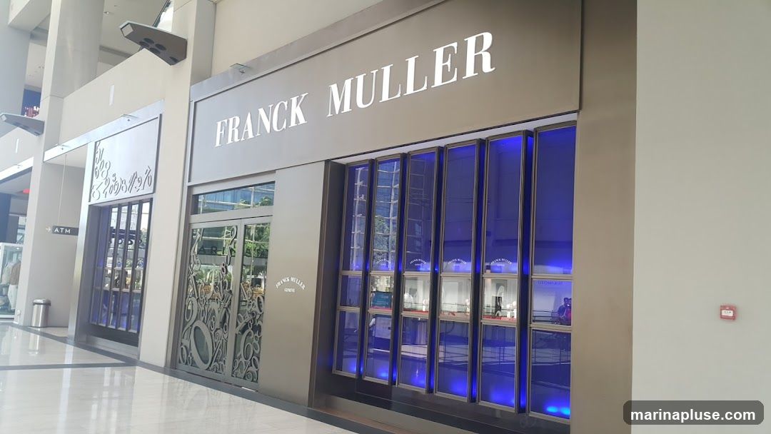The exterior of a Franck Muller watch store. The store has a large, illuminated sign that reads "FRANCK MULLER" above a display window. The display window is divided into multiple sections, each showcasing watches. The store also features decorative metalwork on its entrance doors. To the left of the store, an ATM sign is visible.