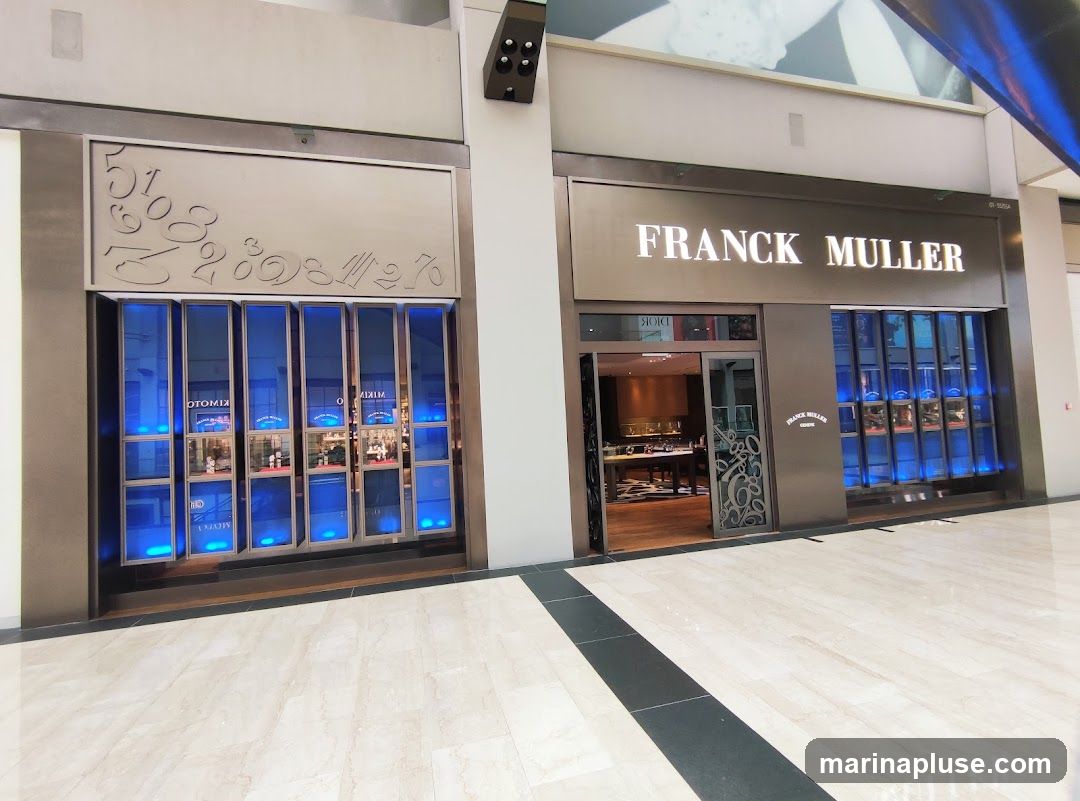 The exterior of a Franck Muller watch store. The store has a large illuminated sign that reads "FRANCK MULLER". To the left of the entrance, there is a display window with multiple compartments, each illuminated with blue light and showcasing watches. To the right of the entrance, there is another similar display window. The entrance itself is a glass door leading into a well-lit interior with display cases and furniture. The floor is tiled with a marble pattern.