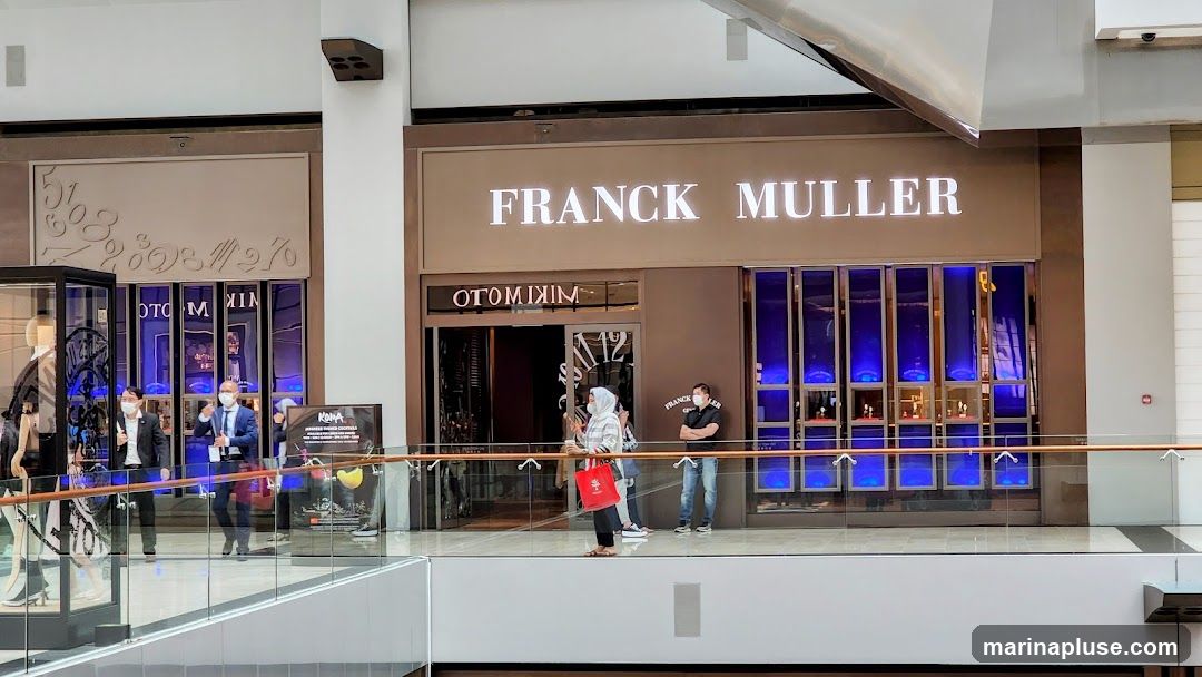 The exterior of a Franck Muller watch store in a mall. The store has a large illuminated sign that reads "FRANCK MULLER" above the entrance. The entrance has mirrored doors and windows displaying watches. Several people are visible in front of the store and on the walkway. To the left of the entrance, there is a display case with mannequins. To the right of the entrance, there is a poster advertising cocktails.