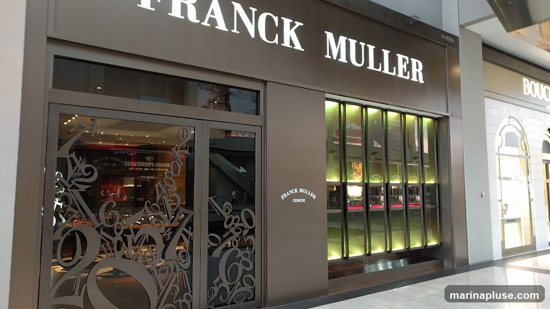 The exterior of a Franck Muller watch store. The store has a large, illuminated sign above the entrance that reads "FRANCK MULLER". The storefront features a glass door with decorative numbers and a display window showcasing watches. To the right of the entrance, there is a vertical display case with multiple illuminated sections, also showing watches.