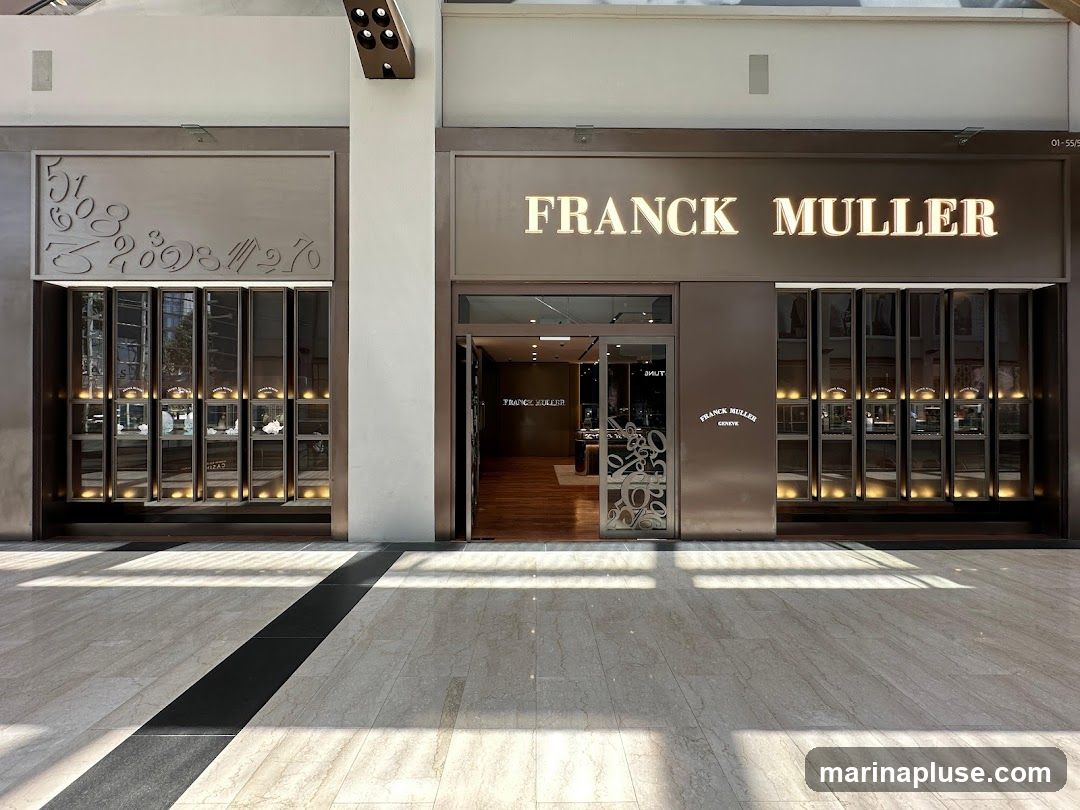 The exterior of a Franck Muller watch store. The store has a large illuminated sign that reads "FRANCK MULLER". To the left of the entrance, there is a display window with numbers and watch cases. To the right of the entrance, there are more display windows showcasing watches. The store entrance is open, revealing the interior.