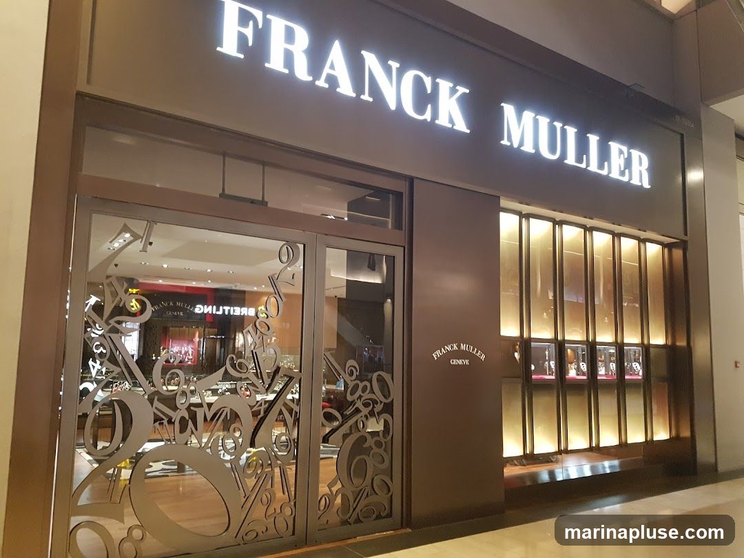 The exterior of a Franck Muller watch store. The store has a large illuminated sign that reads "FRANCK MULLER" above the entrance. The entrance features glass doors with a decorative pattern of numbers. To the right of the entrance, there is a display window with vertical golden panels, showcasing watches inside. A smaller "FRANCK MULLER GENEVE" logo is visible on the wall to the left of the display window.