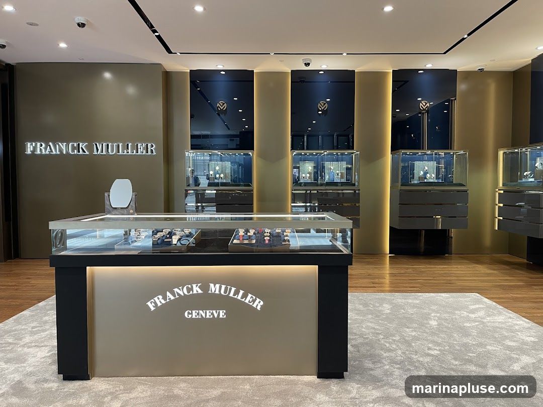 The interior of a Franck Muller watch store, featuring display cases filled with watches. The store has a luxurious and modern design with gold and dark blue accents. A large "FRANCK MULLER" sign is visible on the wall, and the brand name is also illuminated on a central display counter.