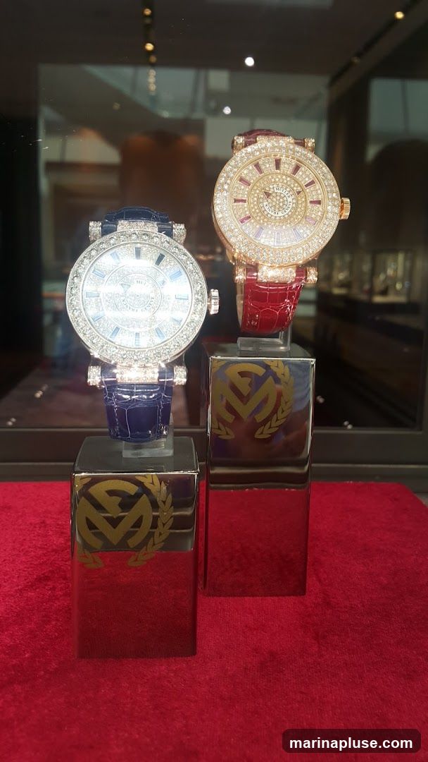 Two luxurious, diamond-encrusted watches are displayed on polished chrome stands against a red velvet background. The watch on the left has a blue alligator strap and a white dial, while the watch on the right has a red alligator strap and a gold-toned dial with ruby accents. Both watches are heavily adorned with diamonds. The chrome stands feature a golden emblem with a laurel wreath. The background appears to be a display case in a jewelry store, with reflections of lights and other merchandise visible.