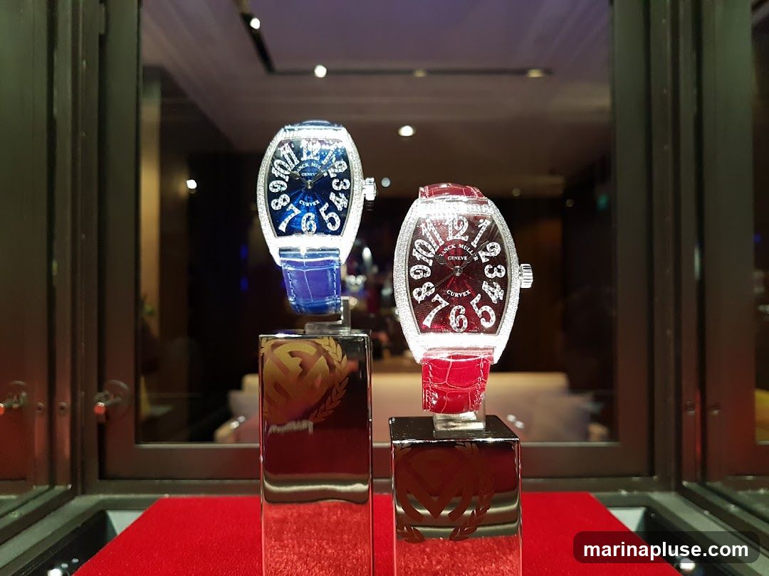 Two Franck Muller watches are displayed on stands in a glass case. The watch on the left has a blue face and a blue strap, and is encrusted with diamonds. The watch on the right has a dark red face and a red strap, and is also encrusted with diamonds. Both watches have the Franck Muller logo and the word "CURVEX" visible on their faces. The stands are reflective and have a logo etched into them. The background is blurred and appears to be an interior of a store.