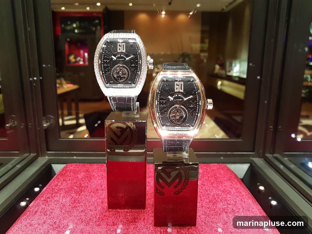 Two Franck Muller watches are displayed on red velvet in a glass case. The watch on the left is encrusted with diamonds around the bezel, while the watch on the right has a rose gold bezel. Both watches have black dials with Roman numerals and a tourbillon complication. The watches are displayed on polished black stands with the Franck Muller logo. The background shows a blurred interior of a luxury store.