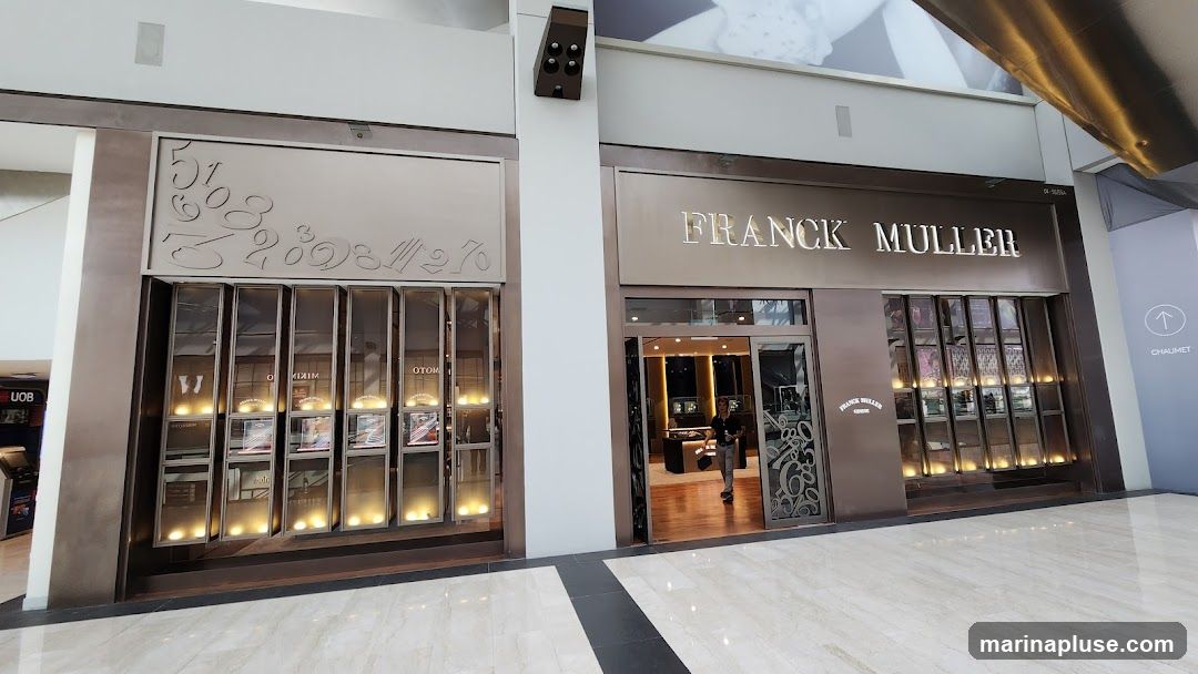 The exterior of a Franck Muller watch store. The store has a dark metallic facade with the brand name "FRANCK MULLER" prominently displayed above the entrance. The storefront features multiple display windows showcasing watches, illuminated from below. To the left of the main entrance, there is another section with display cases and decorative numbers on the wall. A person is visible entering the store.