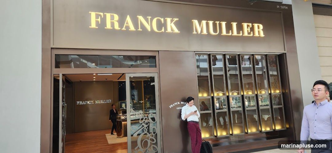 The exterior of a Franck Muller watch store. The store has a large, illuminated sign above the entrance that reads "FRANCK MULLER". The storefront features a glass entrance and display windows showcasing watches. A man in a white shirt and maroon pants is leaning against the wall outside the store, looking at his phone. Another man in a light purple shirt walks past the entrance. Inside, a woman in a dark suit is visible behind a counter.