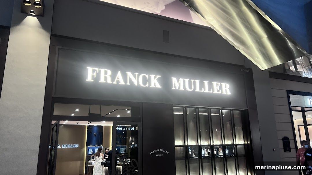 The exterior of a Franck Muller watch store. A large, illuminated sign above the entrance reads "FRANCK MULLER". Below the sign, the store has large glass windows displaying watches and a dark facade. Two people are visible near the entrance, one appearing to be a staff member.