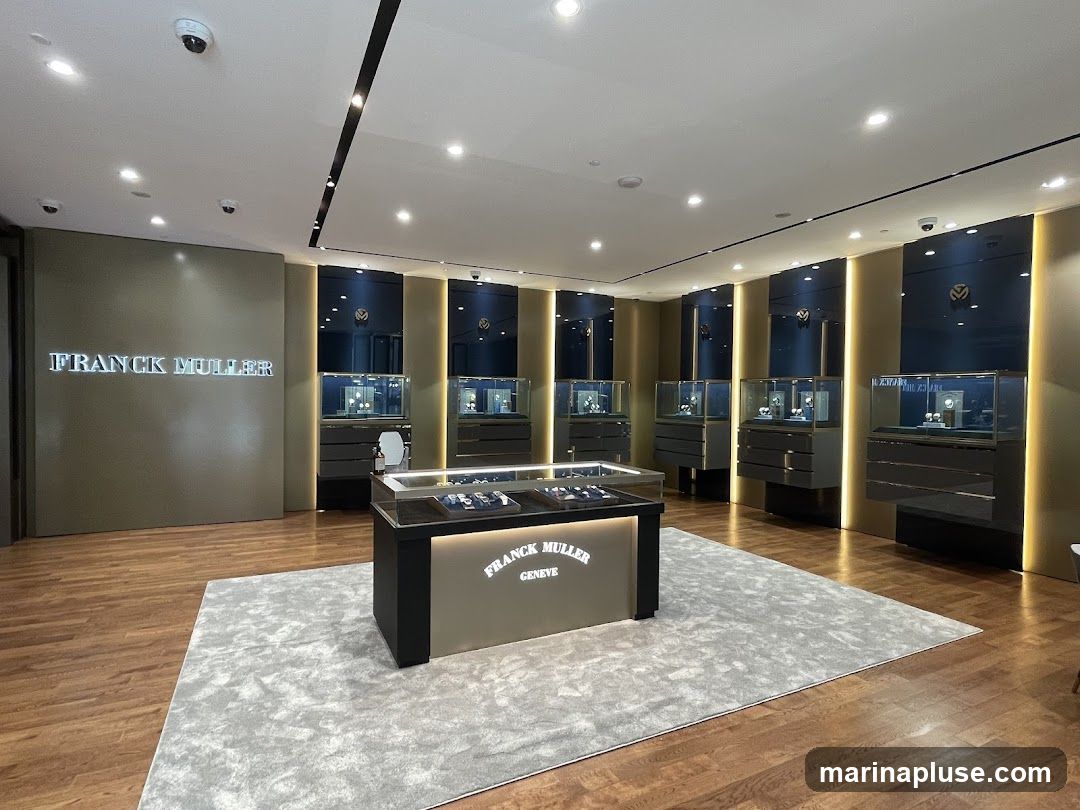 The interior of a Franck Muller watch store, featuring illuminated displays of watches in glass cases. The store has a modern and luxurious design with wooden floors, gold-toned walls, and dark blue accents. The Franck Muller logo is prominently displayed on a wall and on a central display counter.