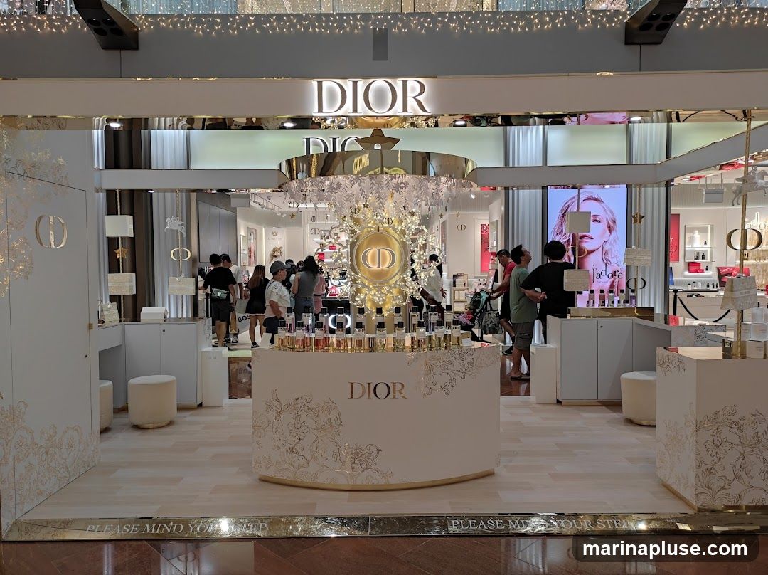 A Dior perfume display is set up in a mall, featuring a central circular display with the Dior logo and numerous perfume bottles. The display is decorated with white and gold ornaments and lights, creating a festive atmosphere. Several people are browsing the products. A large screen displays an advertisement for Dior's J'adore perfume.