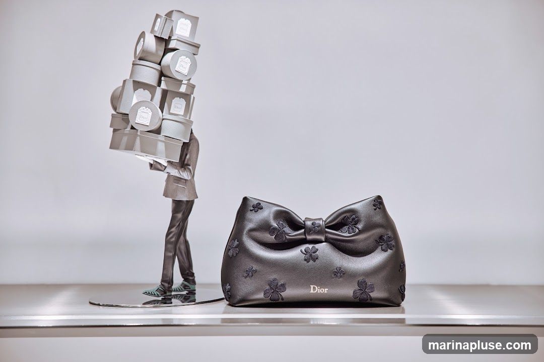 A dark grey Dior clutch bag with a bow detail and embroidered floral motifs is displayed on a reflective surface. To the left, a figurine of a person in a suit carries a tall stack of Dior-branded gift boxes.