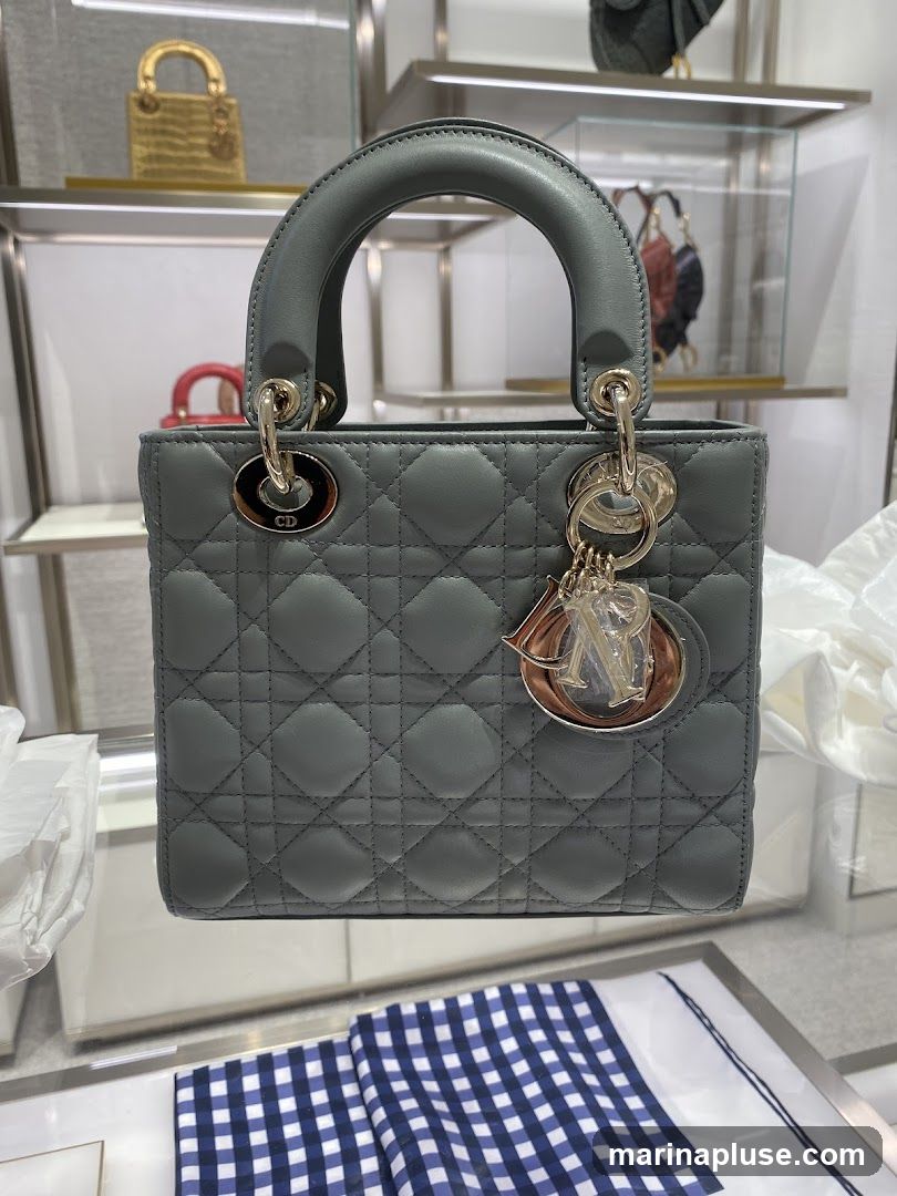 A close-up shot of a grey Dior Lady Dior handbag displayed in a store. The bag features the brand's signature cannage quilting and is adorned with a Dior charm. In the background, other handbags and accessories are visible on shelves.