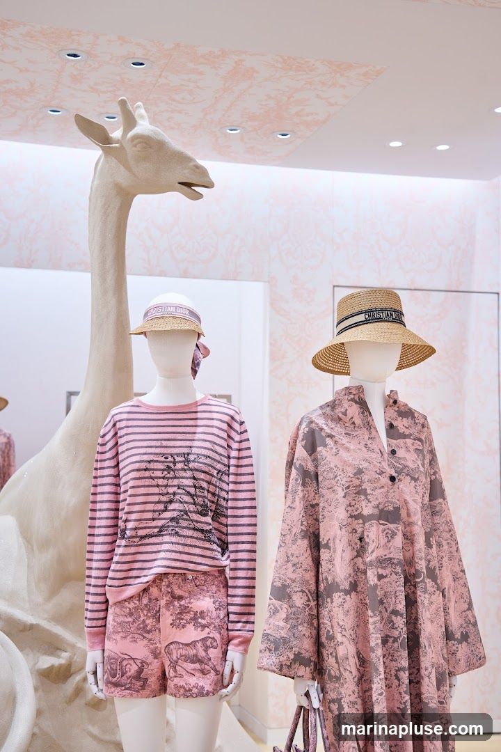 A whimsical display featuring mannequins dressed in pink and grey Dior clothing with animal prints. A large, light-colored giraffe sculpture stands behind the mannequins. The background is adorned with pink wallpaper featuring a toile de Jouy pattern. The mannequins wear straw hats with the "Christian Dior" logo.