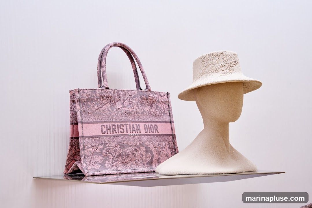 A pink Christian Dior tote bag with a patterned design and the brand name prominently displayed is shown next to a beige mannequin head wearing a textured hat. The items are displayed on a clear shelf against a plain white background.
